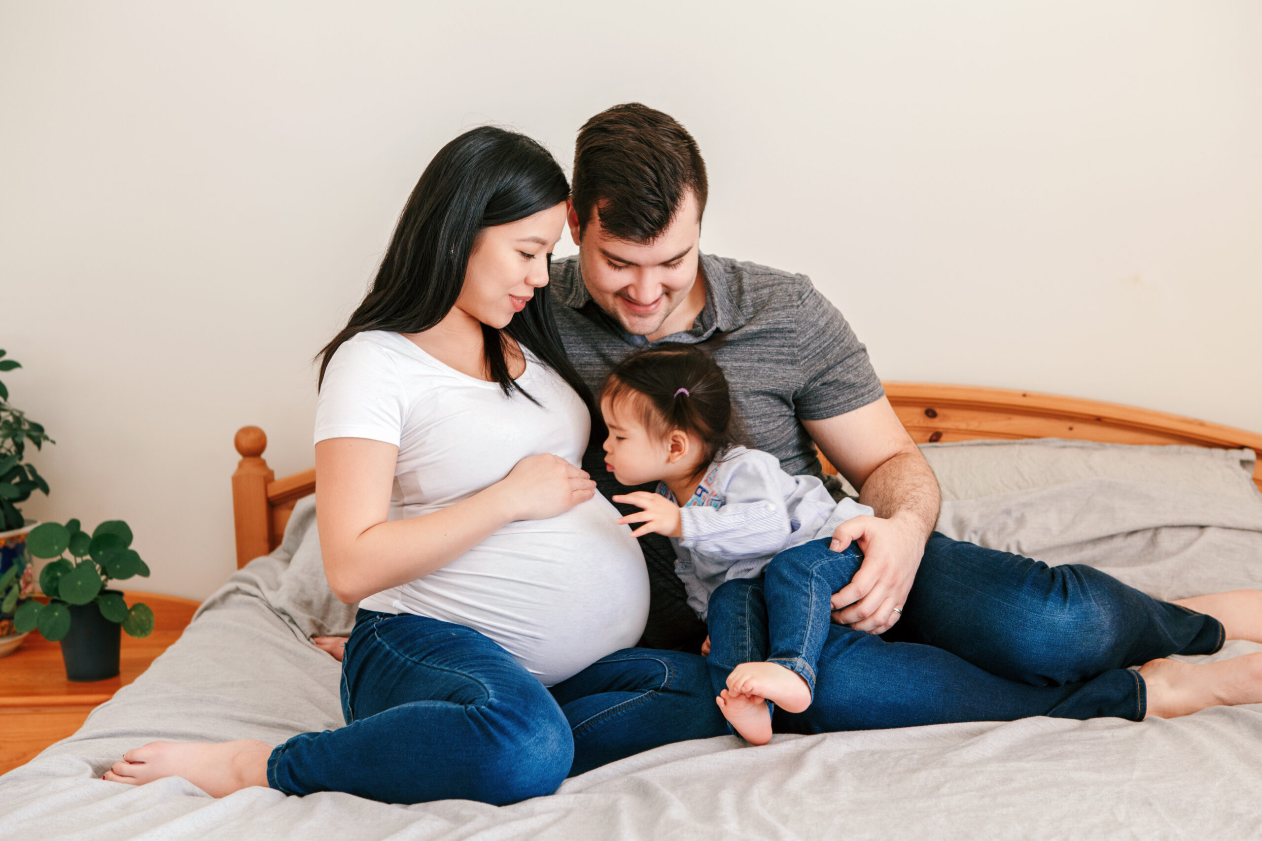 A mix race family sitting on a bed, the child is kissing the pregnant belly