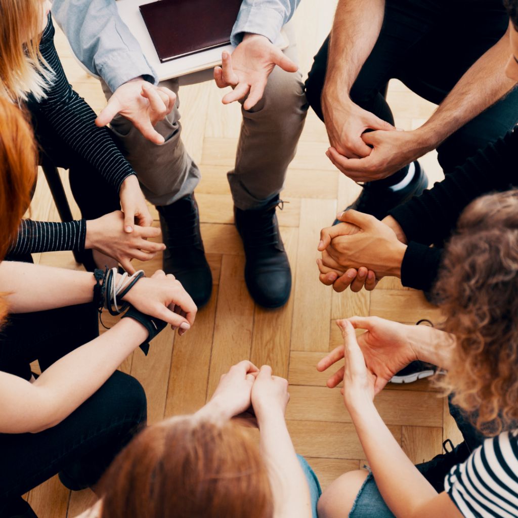 Photo of people sitting in a circle- we mostly see their hands