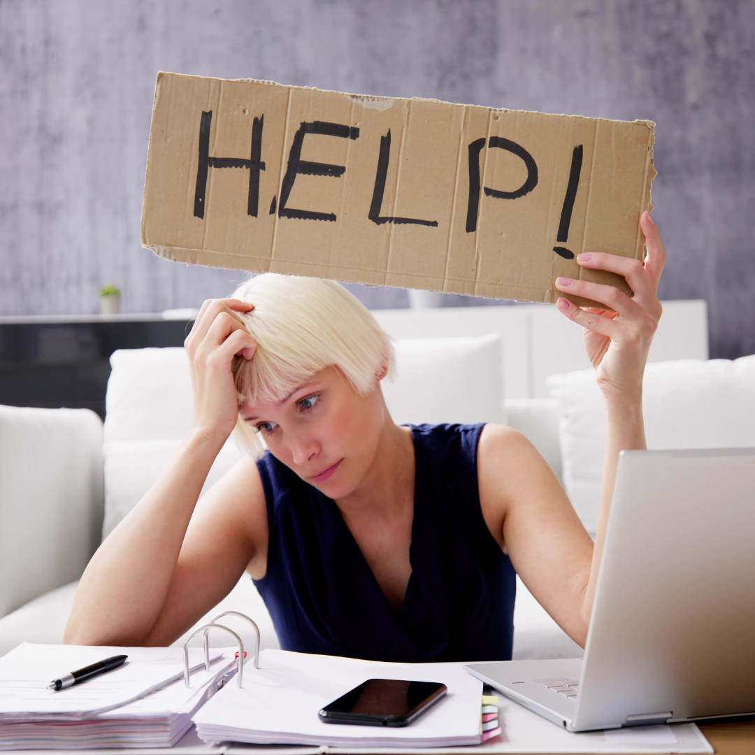 Photo of someone sitting at a desk with computer and paper laid out, and holding a cardboard saying Help