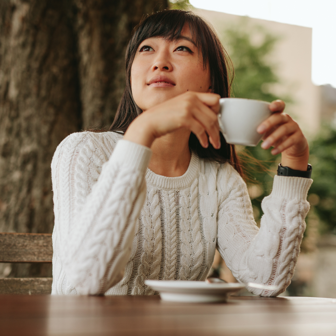 A young person sitting at a table outside and drinking a hot beverage